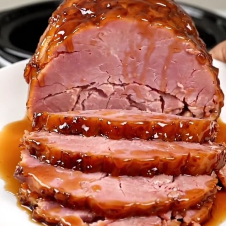 A succulent, glazed boneless ham sitting in a black slow cooker on a white speckled countertop.