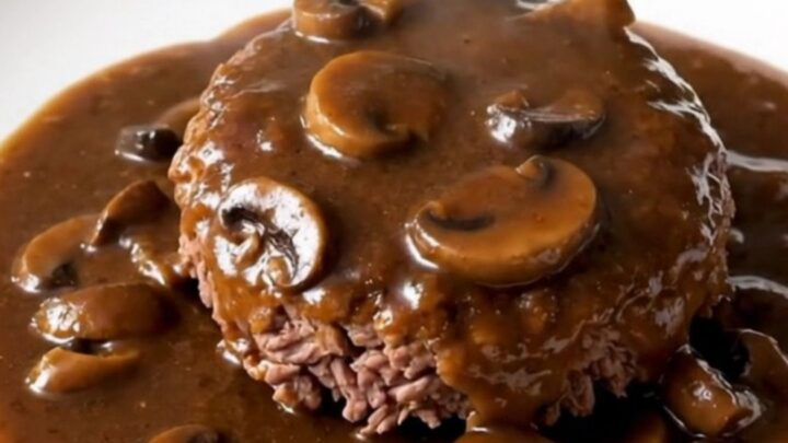 A close-up of a tender Salisbury steak patty covered in a glossy mushroom gravy on a white plate.