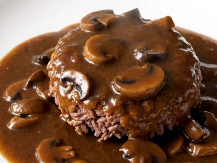 A close-up of a tender Salisbury steak patty covered in a glossy mushroom gravy on a white plate.