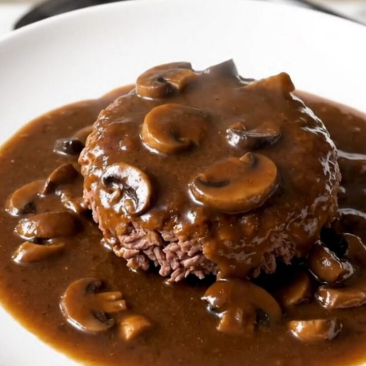 A close-up of a tender Salisbury steak patty covered in a glossy mushroom gravy on a white plate.