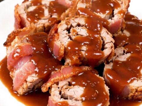 A close-up of slow-cooked bacon-wrapped pork tenderloin slices arranged on a white plate.