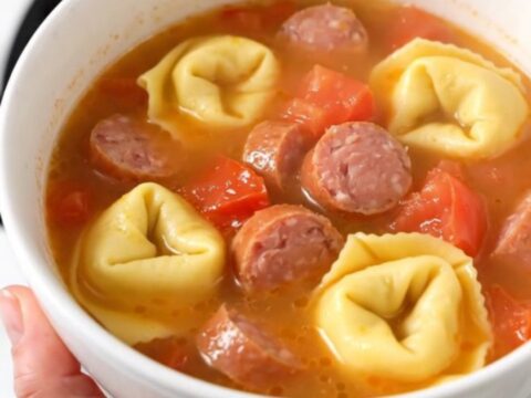 A warm white ceramic bowl filled with creamy sausage and tortellini soup, showing steam rising from the surface.