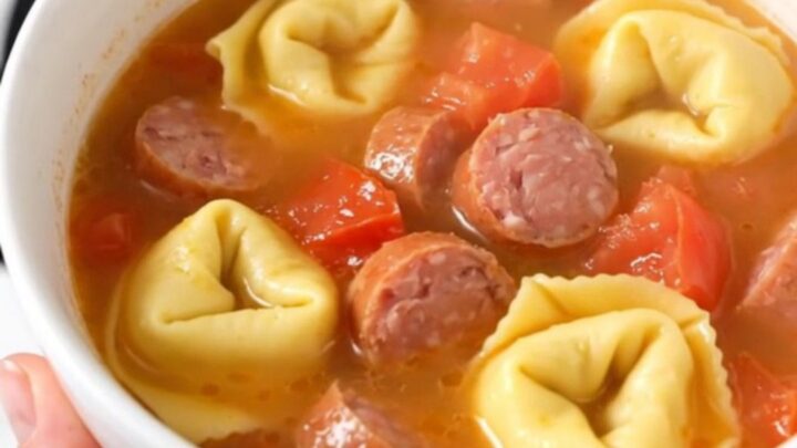 A warm white ceramic bowl filled with creamy sausage and tortellini soup, showing steam rising from the surface.