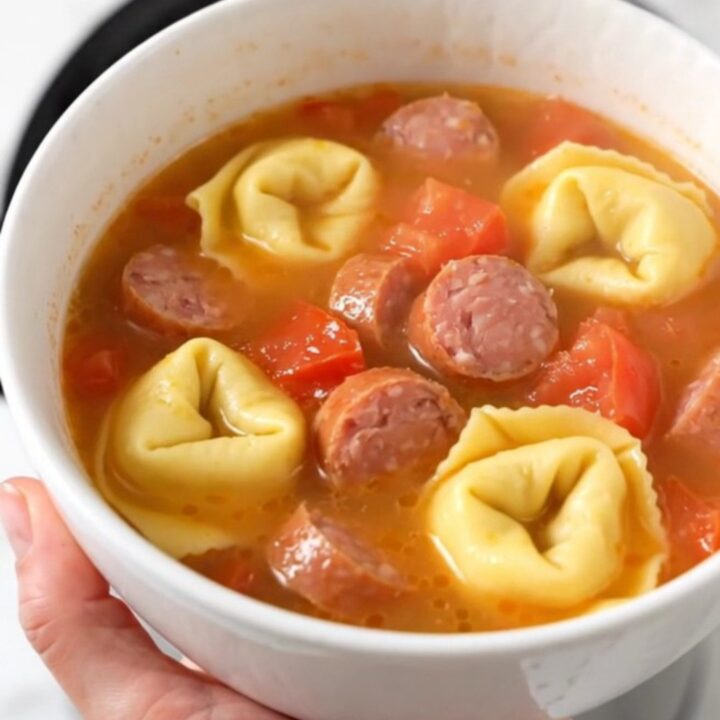 A warm white ceramic bowl filled with creamy sausage and tortellini soup, showing steam rising from the surface.