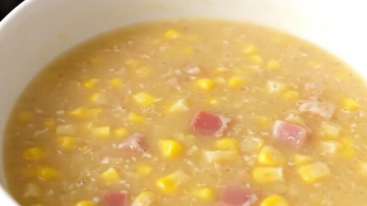 A steaming bowl of creamy corn chowder topped with crispy bacon pieces in a bright kitchen.
