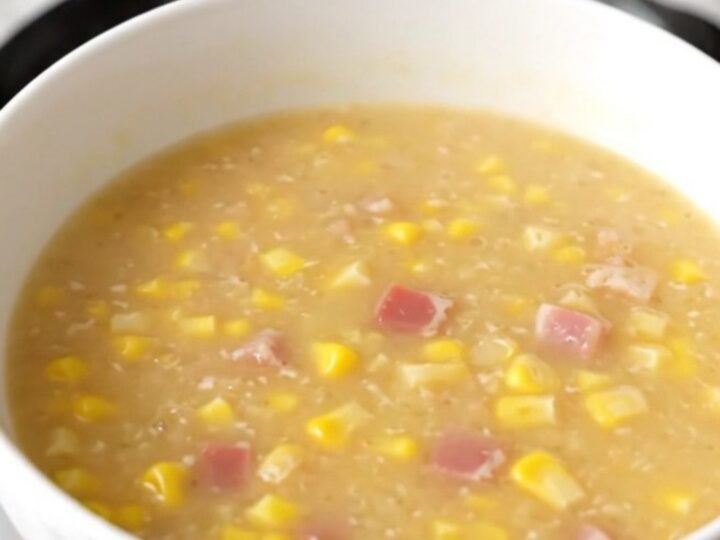 A steaming bowl of creamy corn chowder topped with crispy bacon pieces in a bright kitchen.
