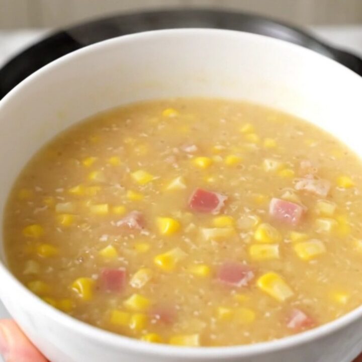 A steaming bowl of creamy corn chowder topped with crispy bacon pieces in a bright kitchen.