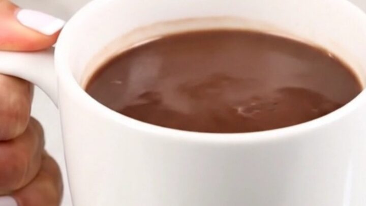 A clear view of creamy, steaming hot chocolate in a white mug against a blurred kitchen background.