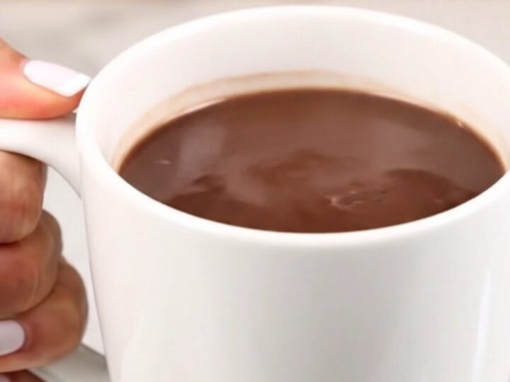 A clear view of creamy, steaming hot chocolate in a white mug against a blurred kitchen background.