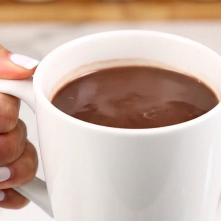 A clear view of creamy, steaming hot chocolate in a white mug against a blurred kitchen background.