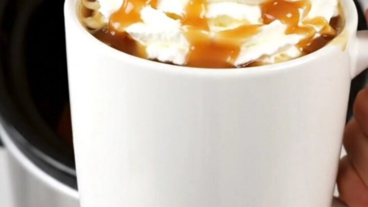 A steaming mug of caramel apple cider topped with whipped cream and a drizzle of caramel on a kitchen counter.