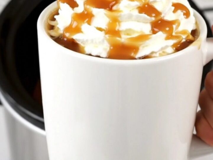 A steaming mug of caramel apple cider topped with whipped cream and a drizzle of caramel on a kitchen counter.