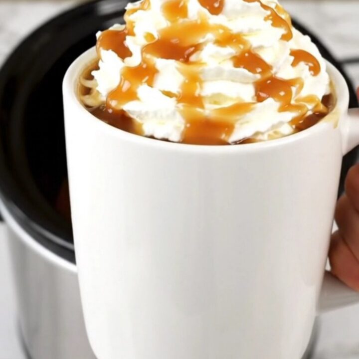 A steaming mug of caramel apple cider topped with whipped cream and a drizzle of caramel on a kitchen counter.