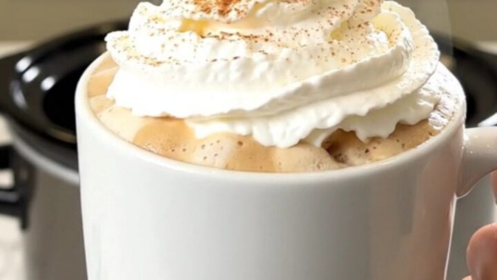 A steaming mug of pumpkin spice latte topped with whipped cream, set against a blurred kitchen background.