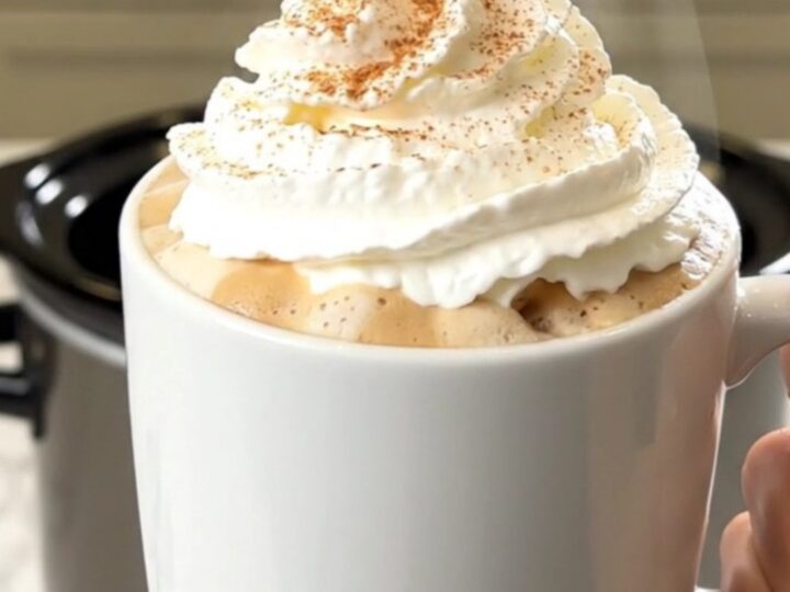 A steaming mug of pumpkin spice latte topped with whipped cream, set against a blurred kitchen background.