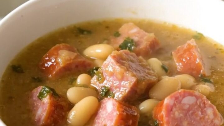 A top-down view of a slow cooker filled with sliced kielbasa, white beans, and seasoned broth before cooking.