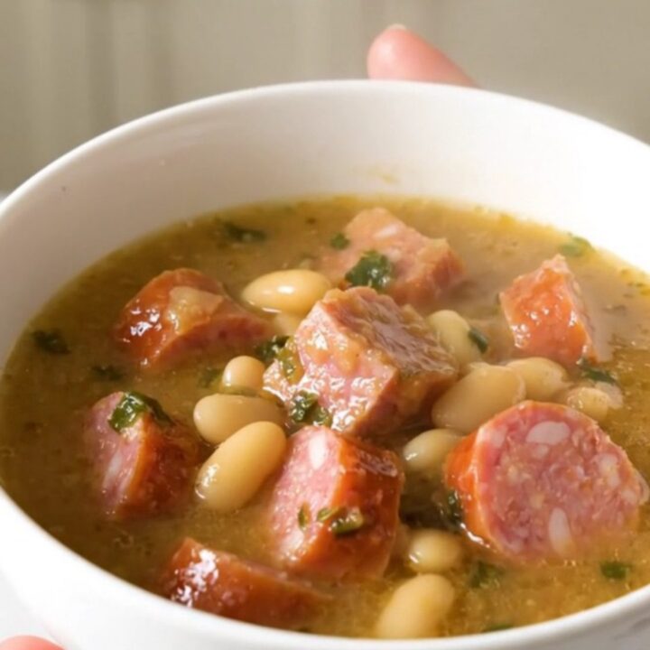 A top-down view of a slow cooker filled with sliced kielbasa, white beans, and seasoned broth before cooking.