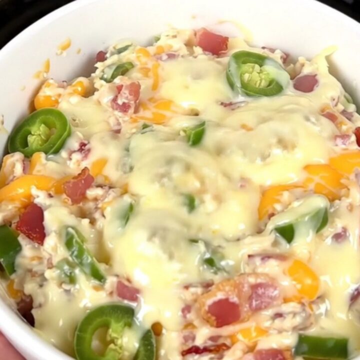 A top-down view of a black slow cooker filled with a creamy, bubbling bacon and corn dip on a white countertop.