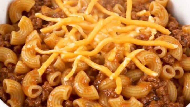 A steaming, glossy beef macaroni goulash in a plain white bowl, showcasing tender pasta and melted cheese.