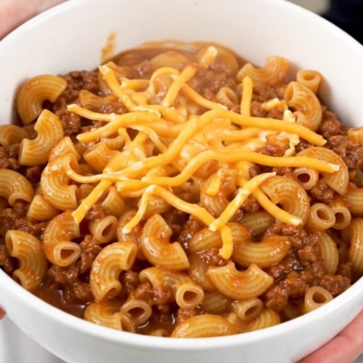 A steaming, glossy beef macaroni goulash in a plain white bowl, showcasing tender pasta and melted cheese.