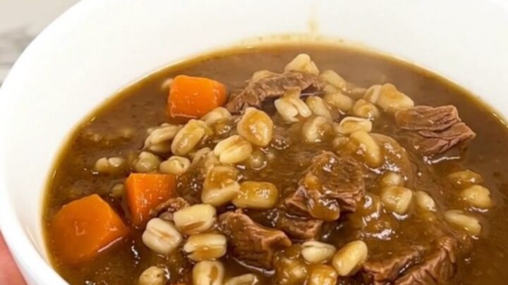 A close overhead shot of a black slow cooker filled with tender beef cubes, bright baby carrots, and a savory, dark broth.