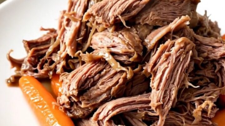 A close-up of a tender, shredded beef chuck roast with baby carrots in a rich, dark sauce.