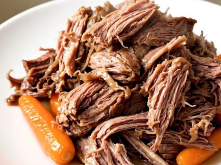 A close-up of a tender, shredded beef chuck roast with baby carrots in a rich, dark sauce.