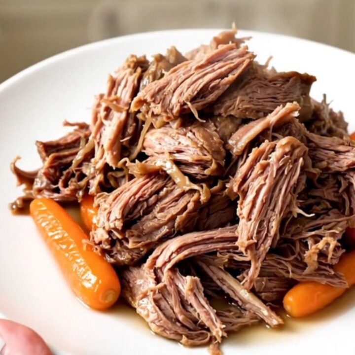 A close-up of a tender, shredded beef chuck roast with baby carrots in a rich, dark sauce.