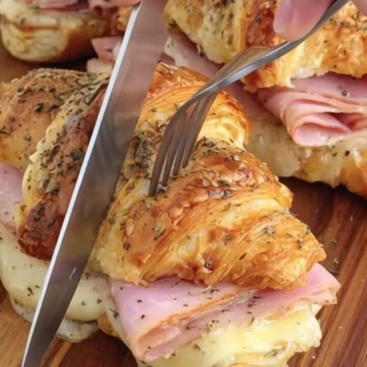 A close-up of a warm, flaky ham and cheese croissant sandwich sliced in half on a wooden board