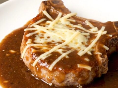 A finished slow cooker French onion pork chop topped with melted Swiss cheese and rich onion gravy on a white plate.