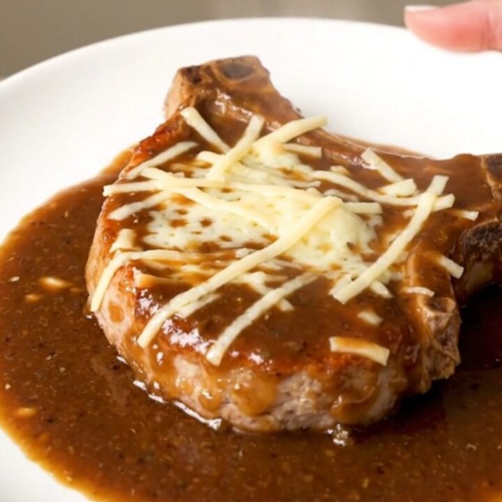 A finished slow cooker French onion pork chop topped with melted Swiss cheese and rich onion gravy on a white plate.
