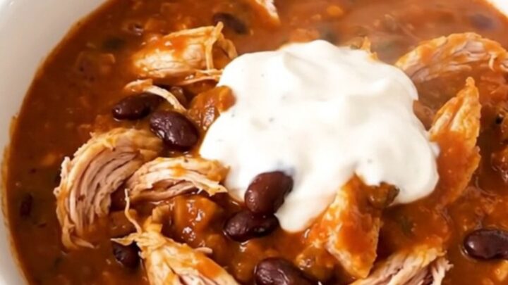 A white bowl filled with glossy, steaming buffalo chicken chili, garnished with a touch of fresh herbs.