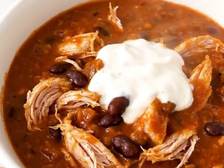 A white bowl filled with glossy, steaming buffalo chicken chili, garnished with a touch of fresh herbs.