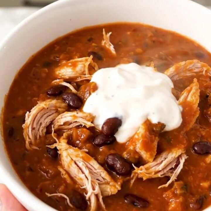 A white bowl filled with glossy, steaming buffalo chicken chili, garnished with a touch of fresh herbs.