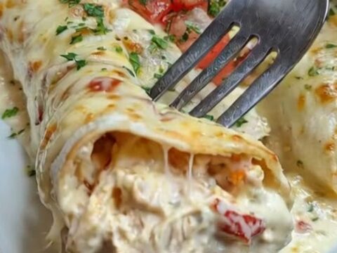 A white dish holding golden, cheesy queso chicken enchiladas garnished with diced tomatoes and fresh cilantro with rising