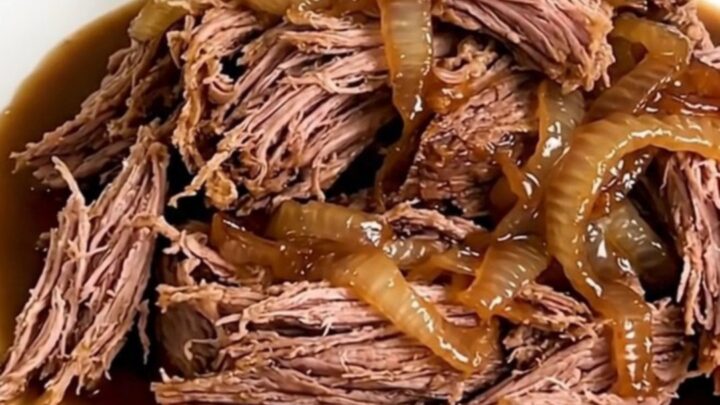 A close-up shot of a tender, shredded beef chuck roast in a slow cooker, featuring a rich barbecue glaze and sweet onions.