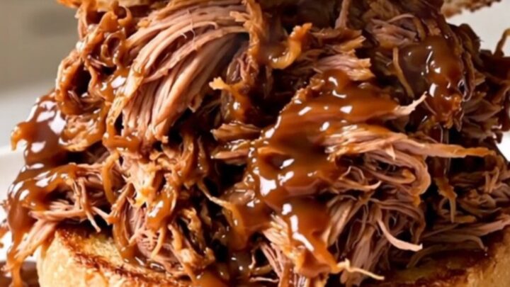 A close-up of a juicy pulled pork sandwich on a toasted brioche bun with a blurred kitchen background.
