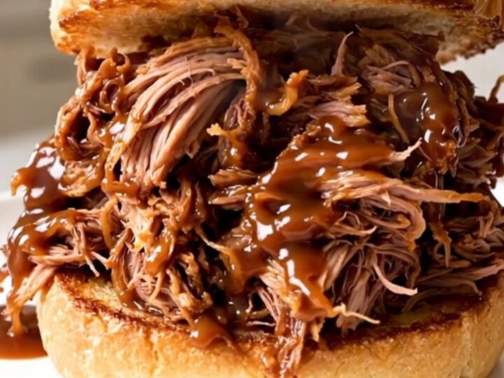 A close-up of a juicy pulled pork sandwich on a toasted brioche bun with a blurred kitchen background.