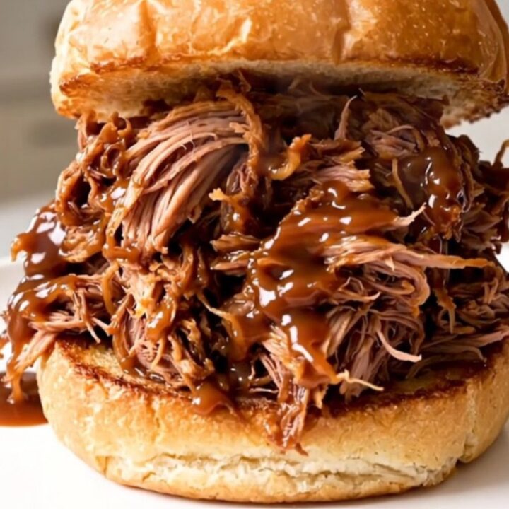 A close-up of a juicy pulled pork sandwich on a toasted brioche bun with a blurred kitchen background.