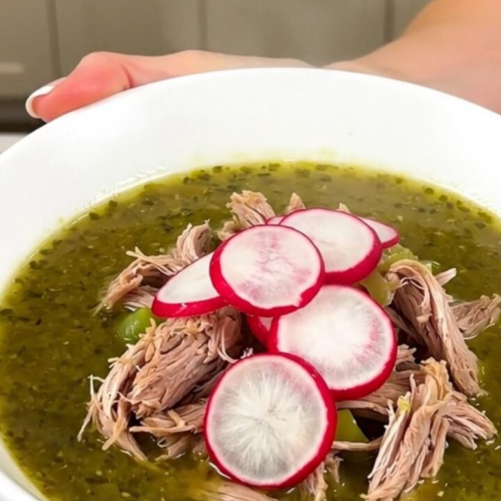 A steaming white bowl filled with shredded pork pozole verde, topped with fresh radish slices.