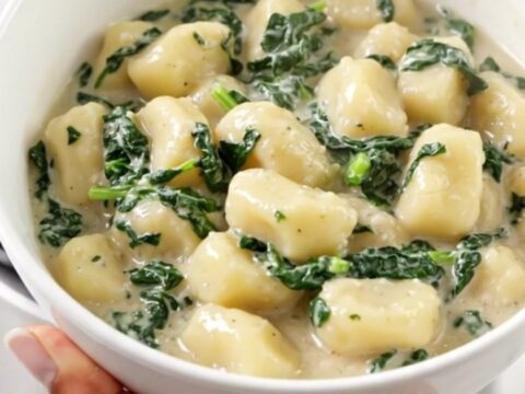 A white bowl filled with creamy spinach and ricotta gnocchi, steam rising from the dish with blurred kitchen cabinets