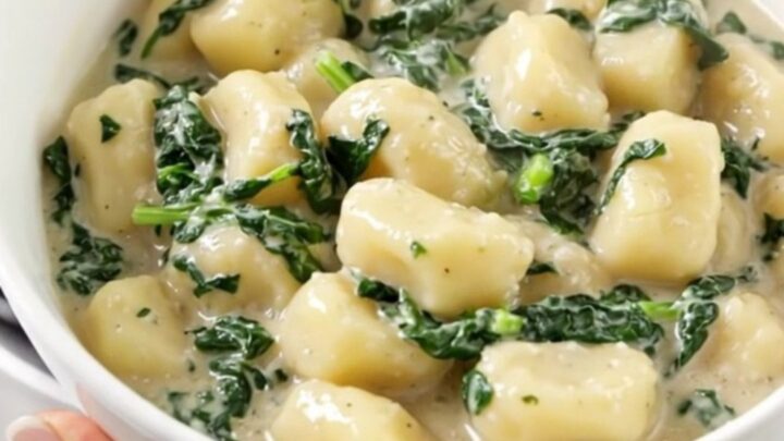 A white bowl filled with creamy spinach and ricotta gnocchi, steam rising from the dish with blurred kitchen cabinets