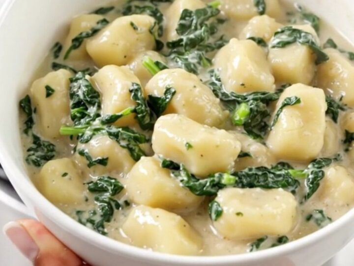 A white bowl filled with creamy spinach and ricotta gnocchi, steam rising from the dish with blurred kitchen cabinets