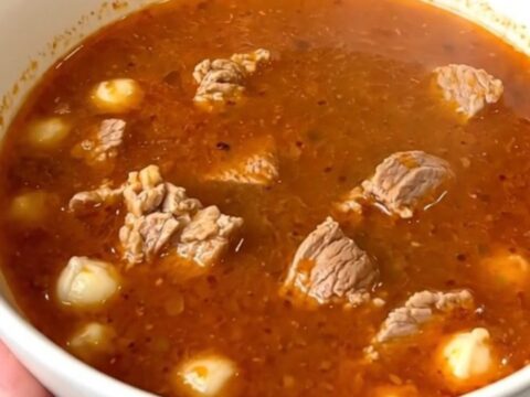 A steaming bowl of pork pozole rojo filled with tender meat and hominy, set against a blurred kitchen background.