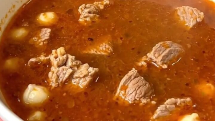 A steaming bowl of pork pozole rojo filled with tender meat and hominy, set against a blurred kitchen background.