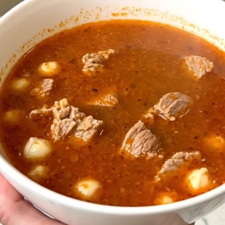 A steaming bowl of pork pozole rojo filled with tender meat and hominy, set against a blurred kitchen background.