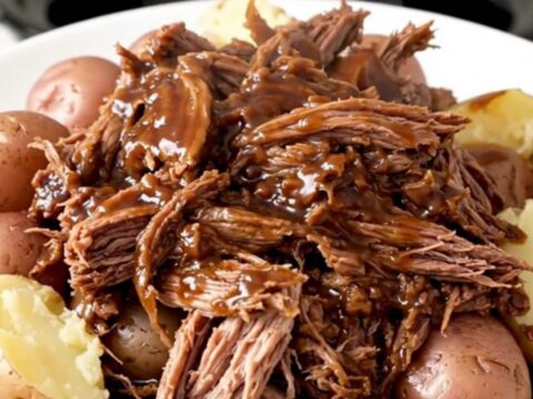 A hearty, rustic slow cooker pot roast served with tender red potatoes and a rich, dark glossy gravy on a white plate.