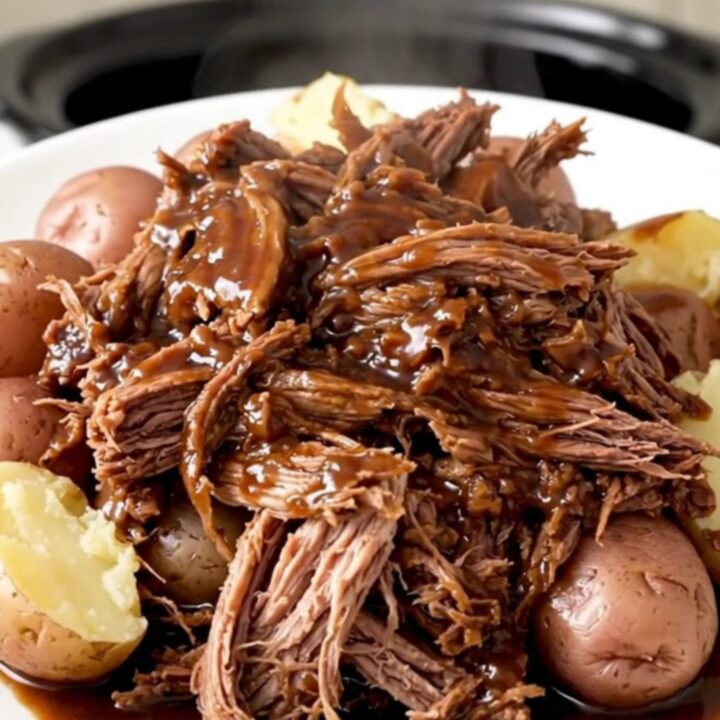 A hearty, rustic slow cooker pot roast served with tender red potatoes and a rich, dark glossy gravy on a white plate.