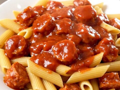 A steaming plate of penne pasta tossed in a rich marinara sauce with sliced Italian sausage and bell peppers.
