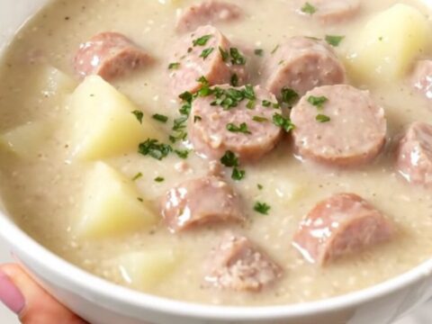 A steaming bowl of creamy sausage and potato chowder served in a white bowl with a blurred kitchen background.